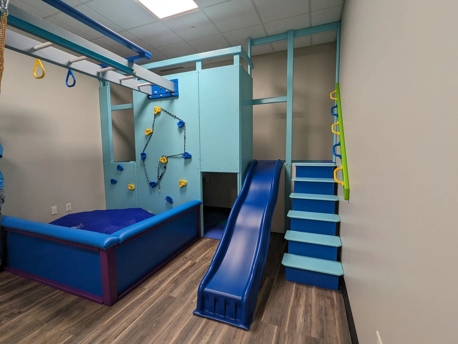 Blue padded ball pit beside a blue slide, teal stairs, and a climbing wall under an overhead ring track