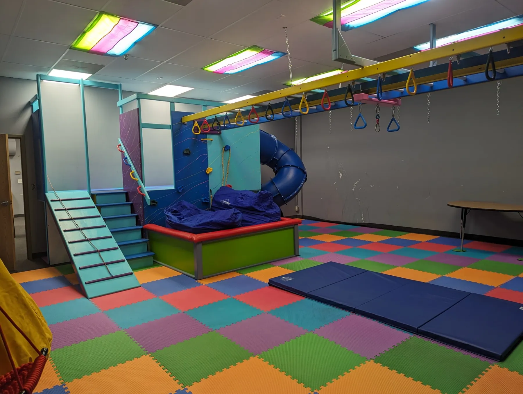 Teal and blue play tower with a slatted ramp, stairs, a climbing wall with holds, a padded ball pit, a blue tube slide, and a yellow overhead ring track
