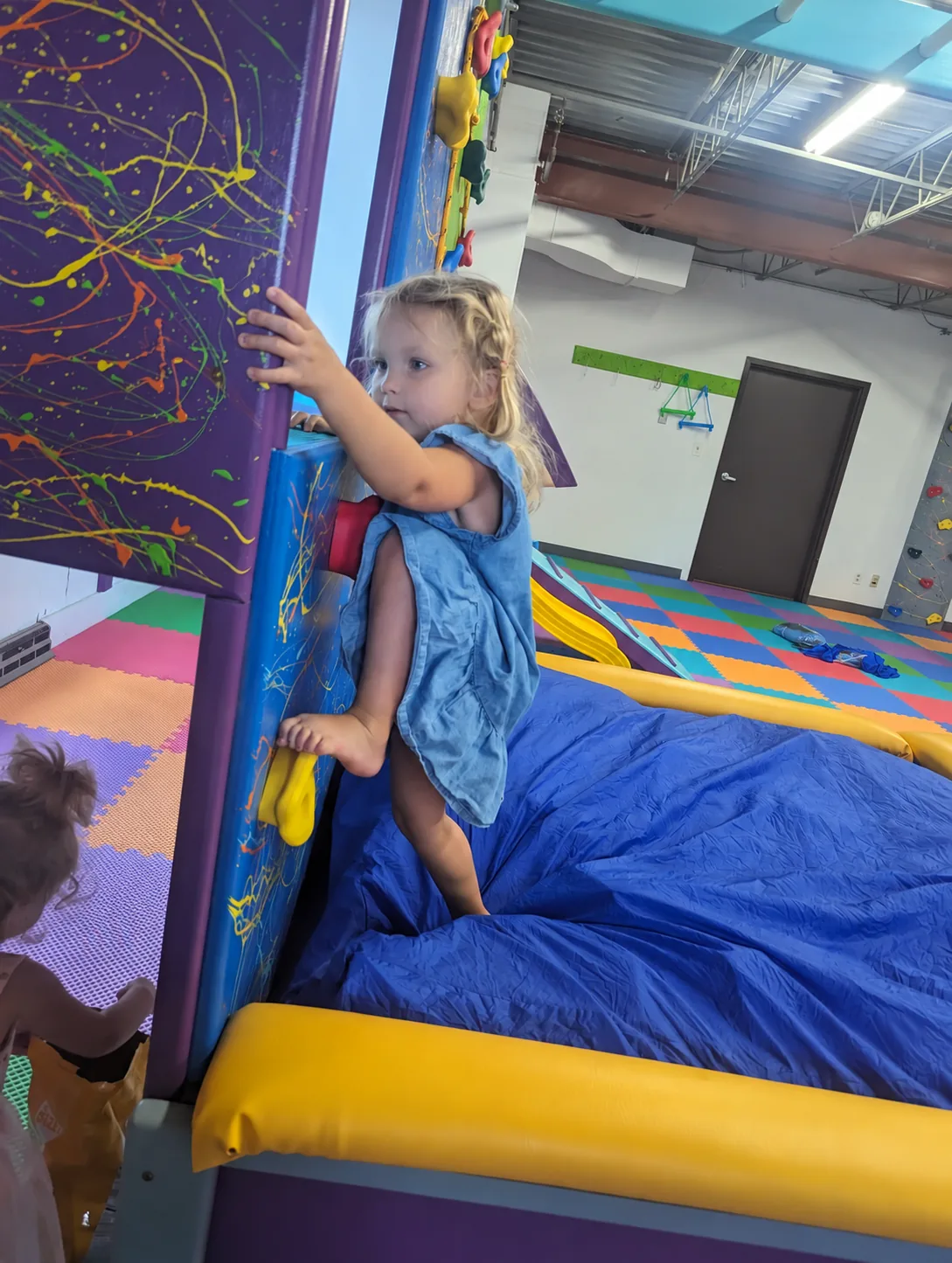 Young child in a blue dress climbing a splatter-painted climbing wall above a padded crash mat in a sensory gym