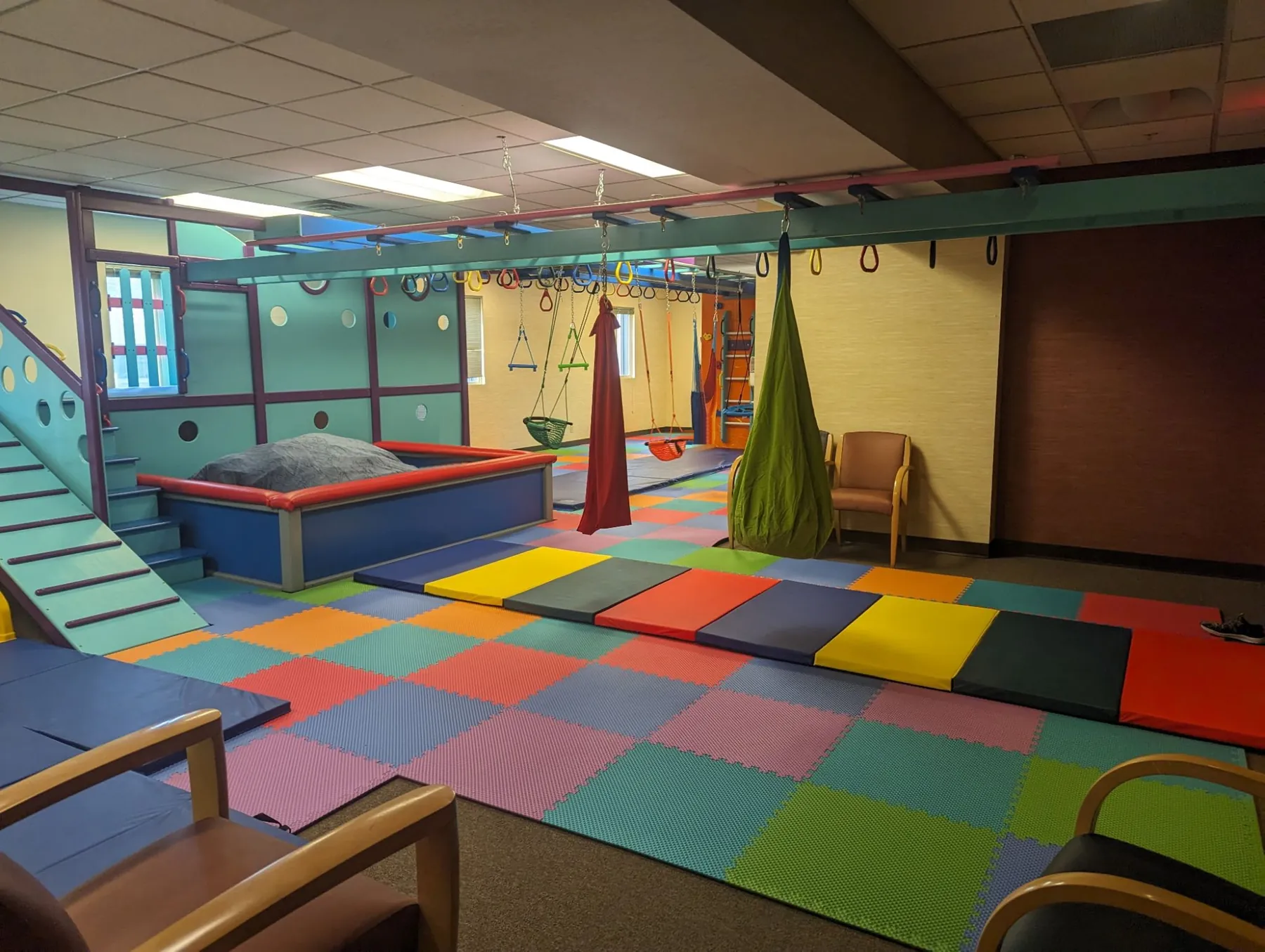 Sensory gym with a padded ball pit beside a teal play structure, an overhead ring track with trapeze rings and hanging swings, and long colorful tumbling mats on foam tiles