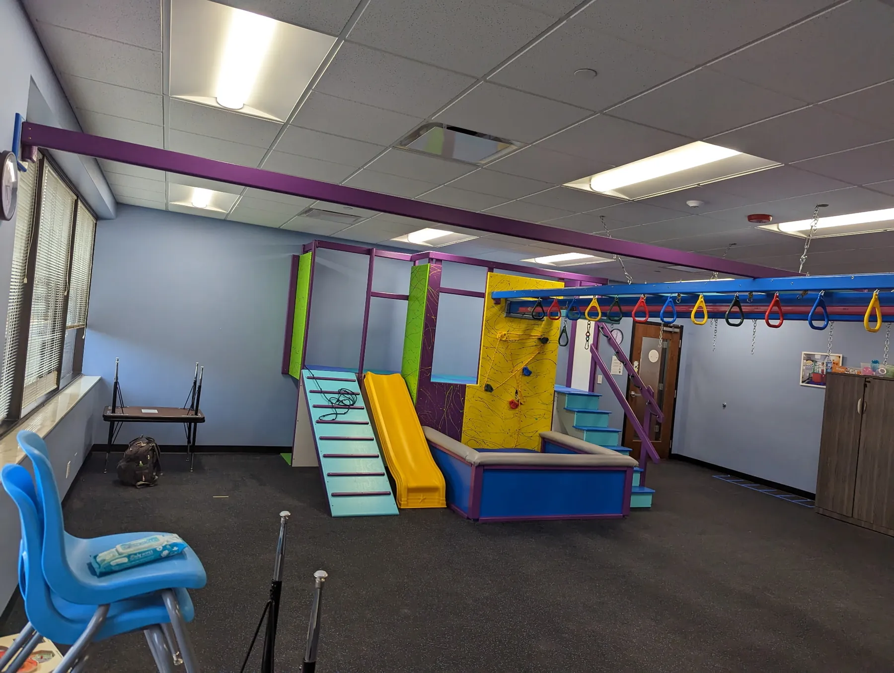 Clinic sensory gym with a slatted ramp, yellow slide, ball pit, stairs, and yellow climbing wall beneath an overhead ring track