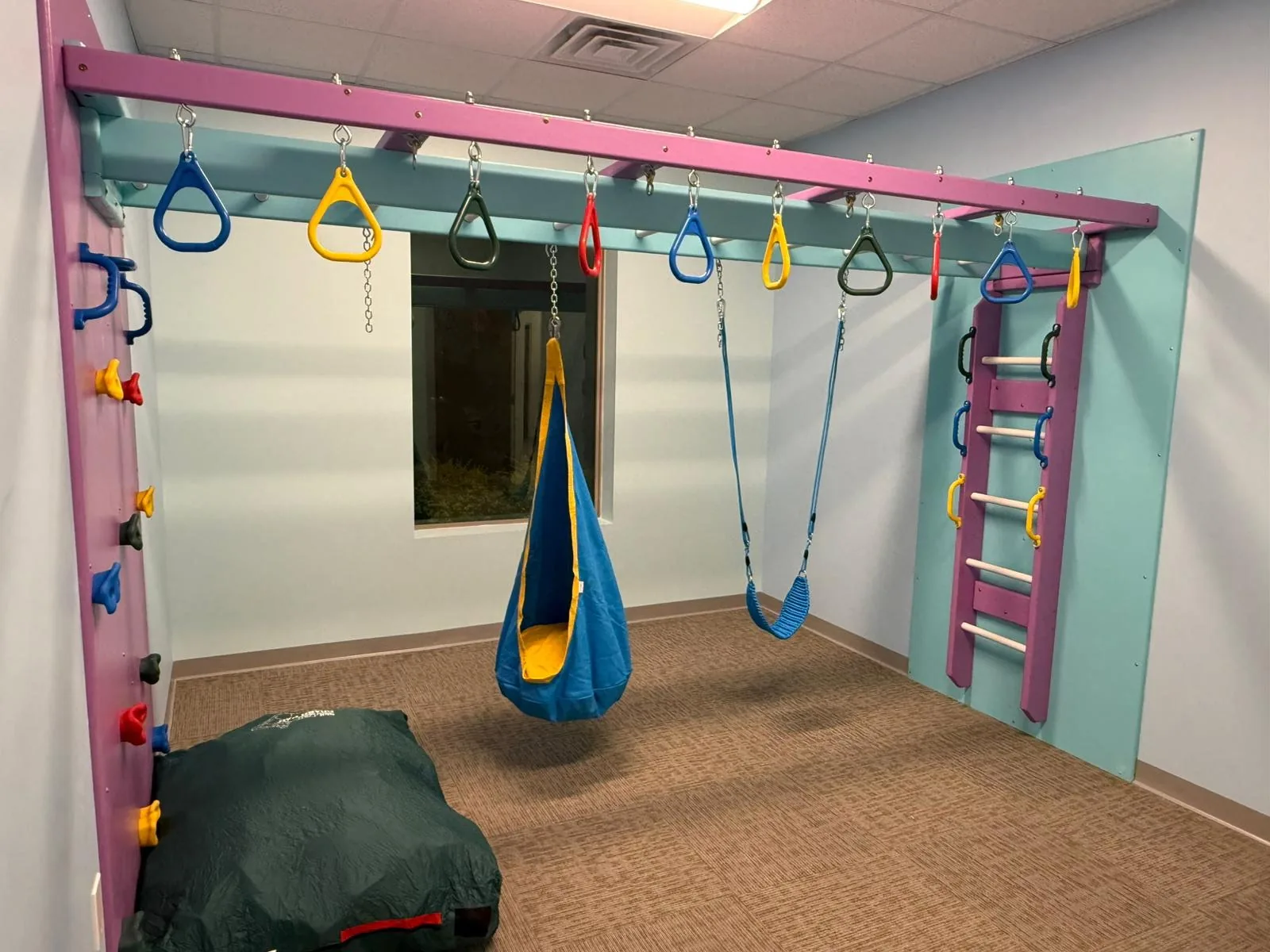 Compact sensory room with a pink and teal frame, trapeze rings, cocoon swing, rope swing, climbing holds, and a wall ladder