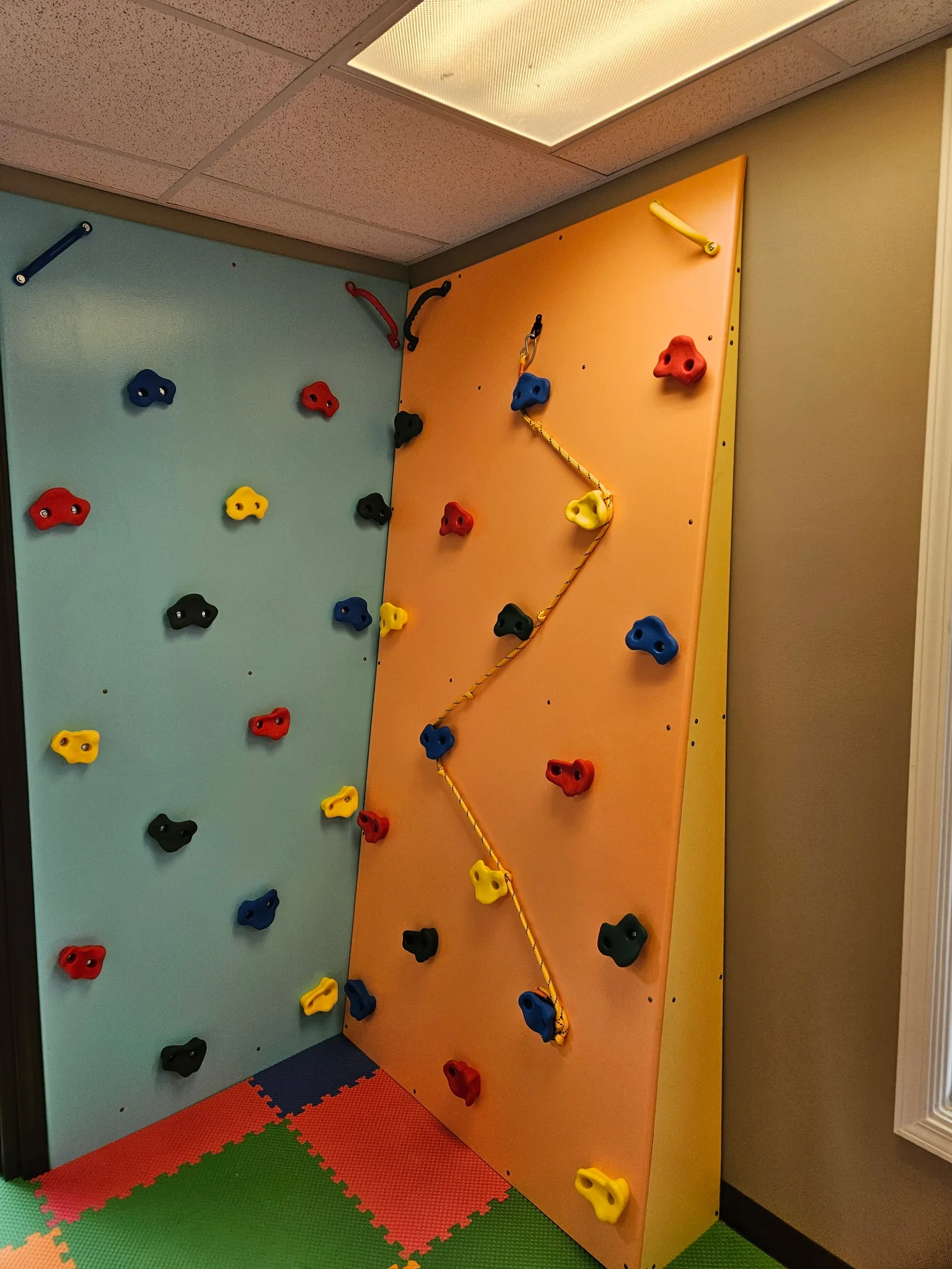 Corner climbing walls in teal and orange with colorful holds and a rope route above foam floor tiles