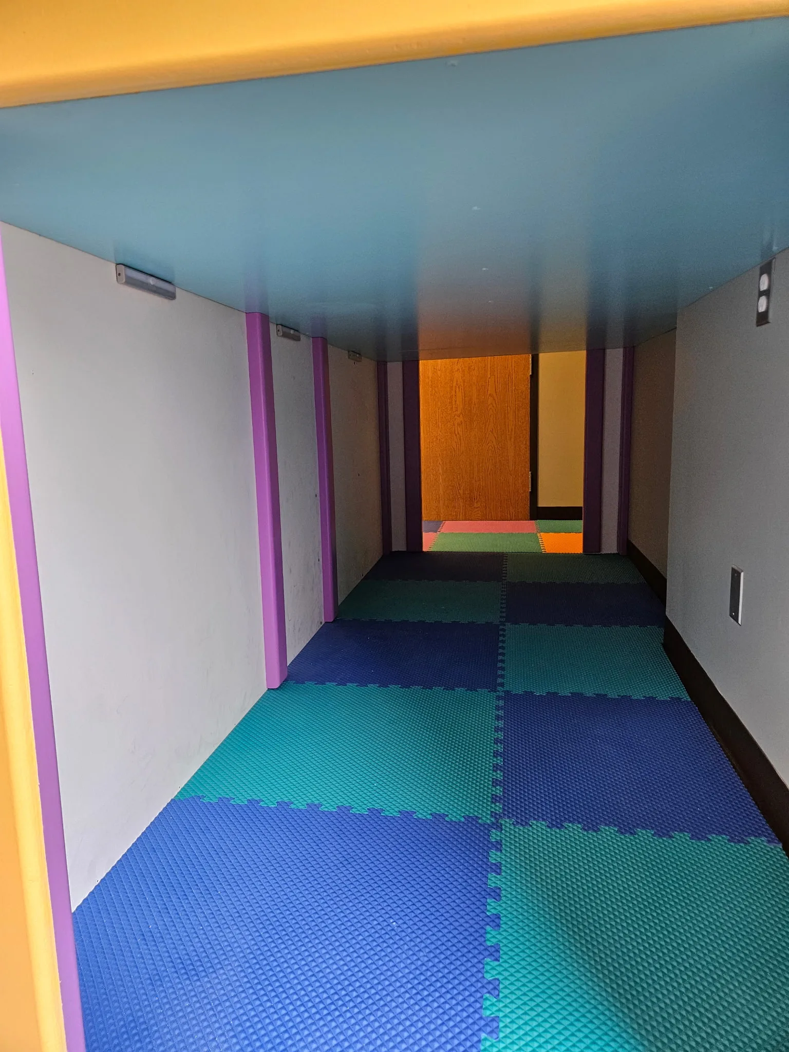 Inside a padded crawl tunnel with blue and teal foam tiles leading to an open doorway