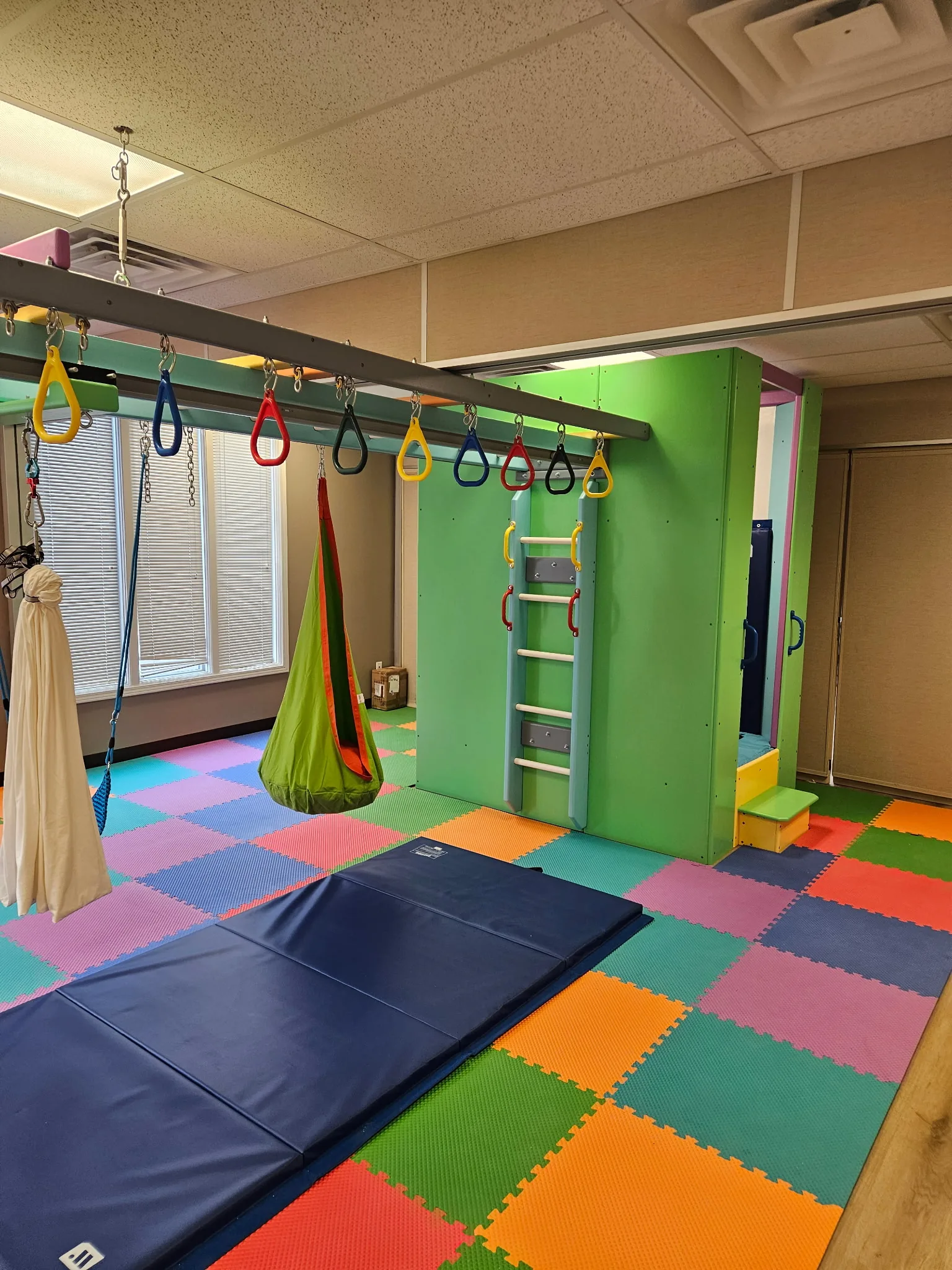 Green climbing tower with a ladder, cocoon swing, trapeze rings, and a blue crash mat on foam tiles