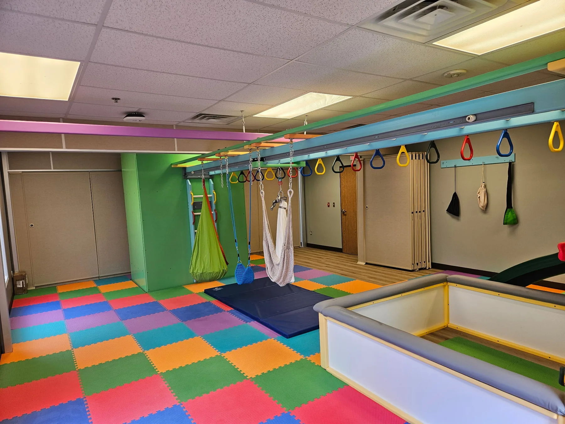 Open sensory gym room with an overhead ring track, suspended swings, a green climbing tower, and colorful foam floor