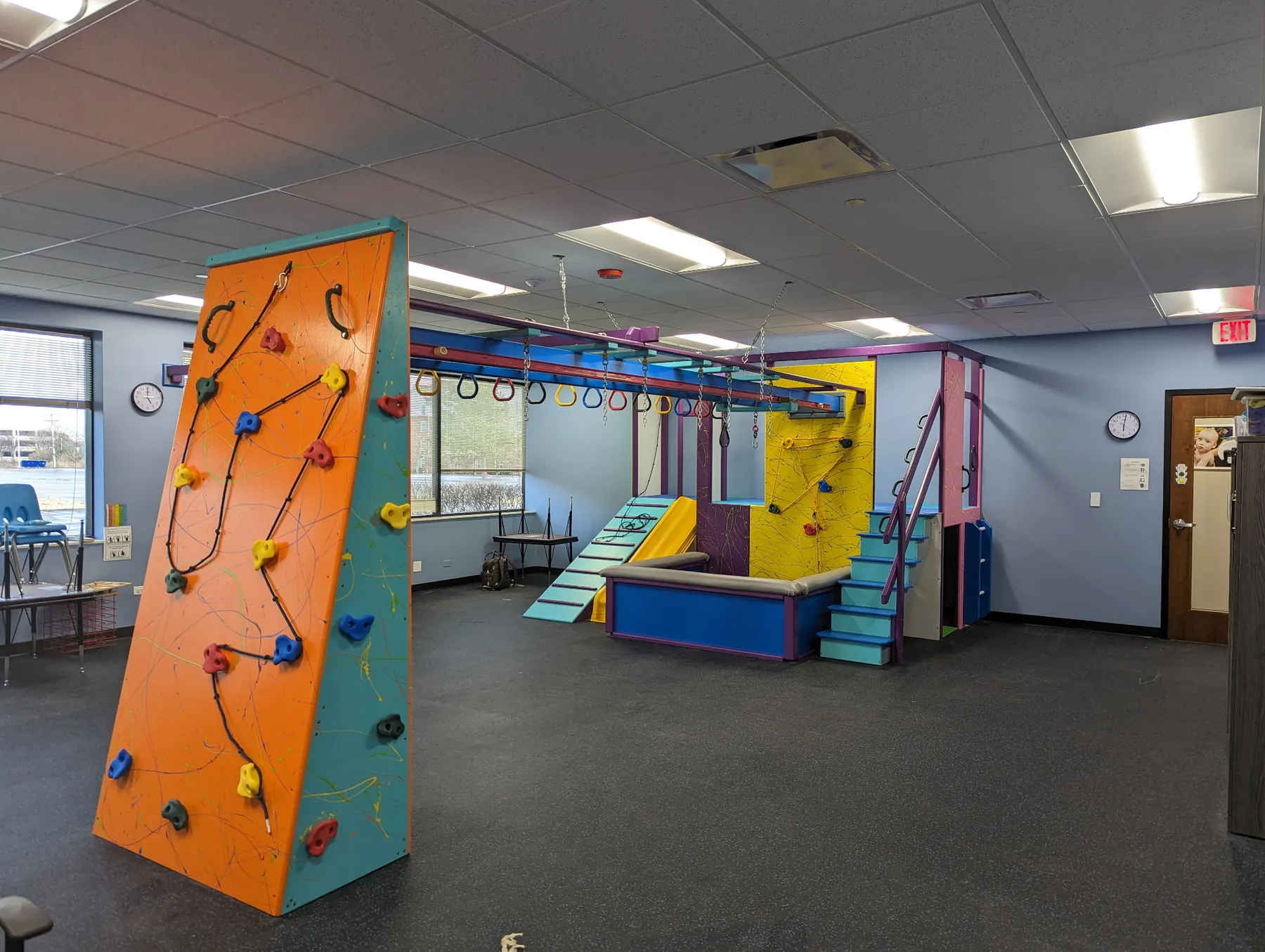 Orange angled climbing wall standing in an open sensory gym with an overhead track, slide, and ball pit behind it