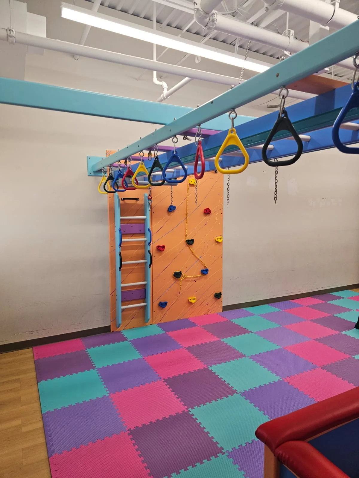 Orange climbing wall with a rope route and a wall ladder beneath an overhead ring track with colorful trapeze rings