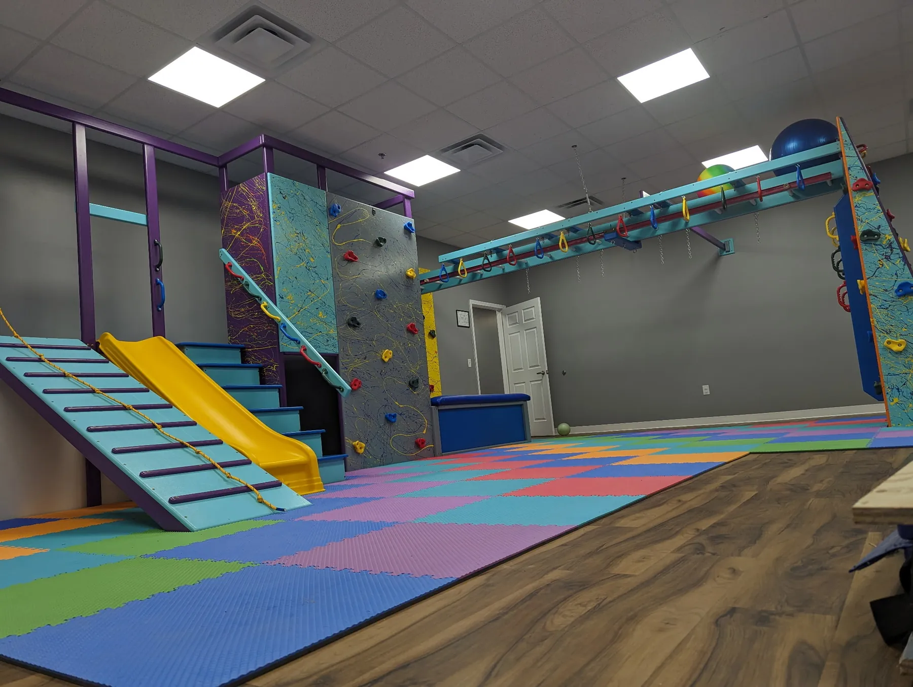 Sensory gym with a slatted ramp, yellow slide, climbing wall, and overhead ring track above foam tiles and a wood floor