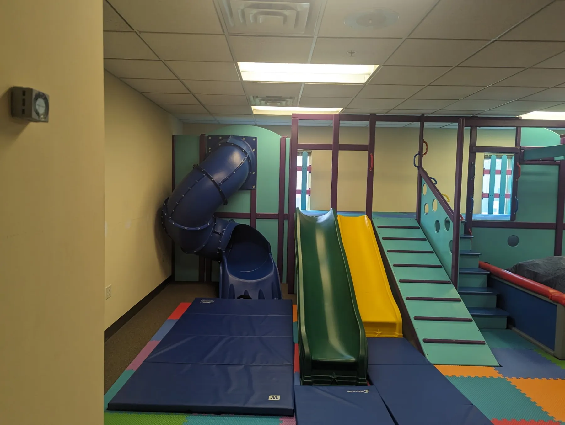 Raised play structure with a blue spiral tube slide, green and yellow side-by-side slides, a slatted climbing ramp, and padded stairs above blue crash mats