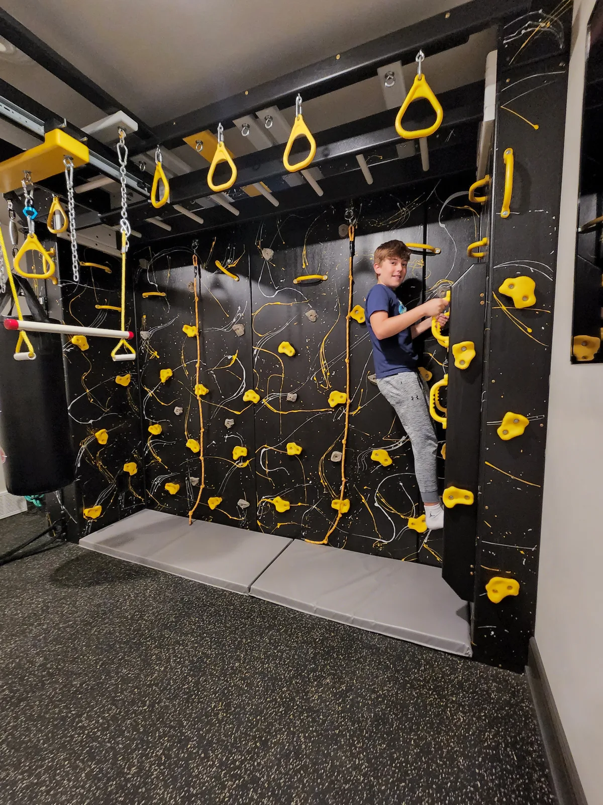 Child climbing a black wall with yellow and green holds beneath yellow overhead bars