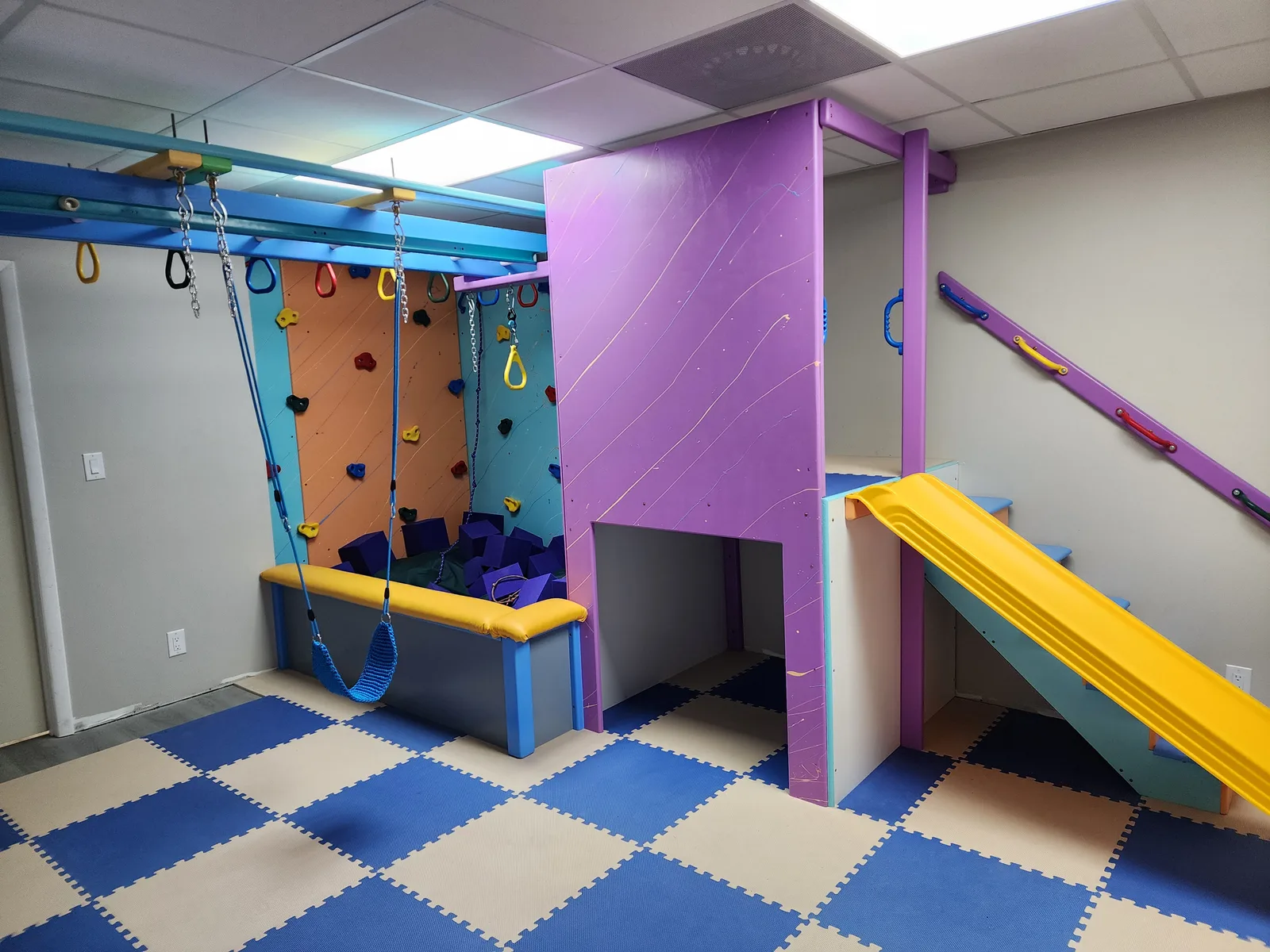 Installed sensory gym with a purple climbing structure, a yellow slide, overhead bars, and a padded landing area