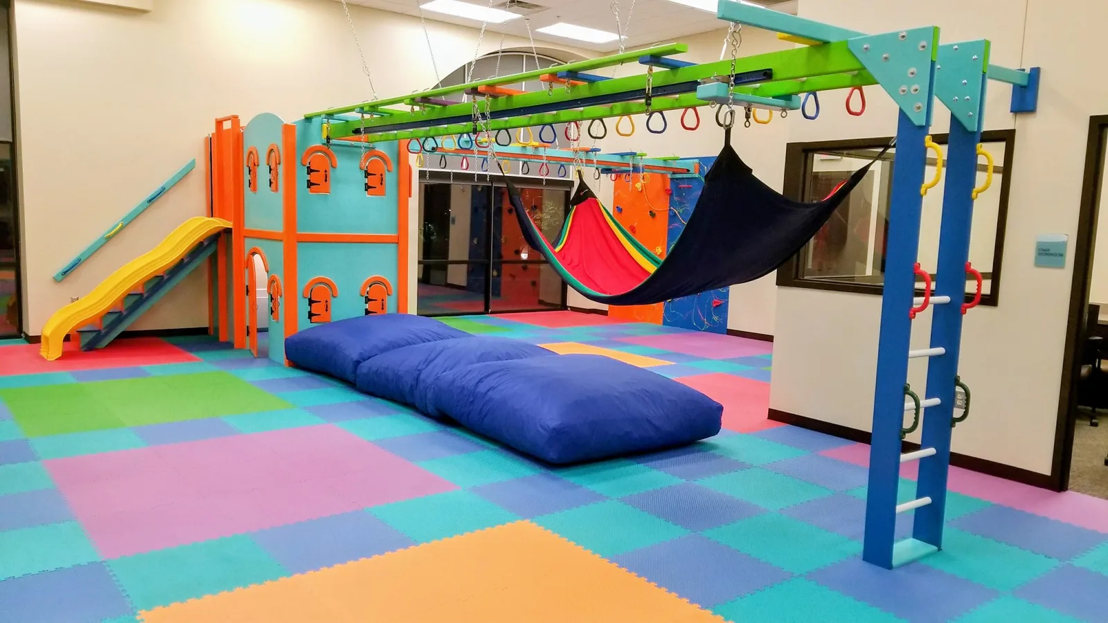 Orange and teal climbing panels, a yellow slide, fabric swings, and blue landing pads over a colorful floor
