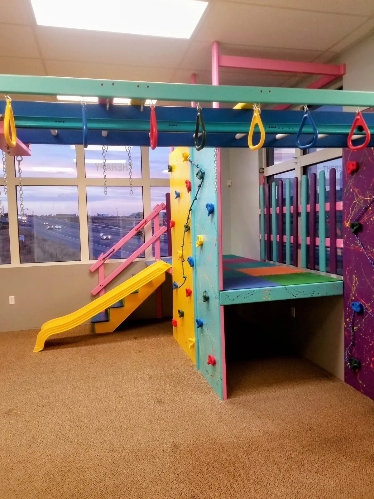Yellow slide and suspended equipment beside large windows in a carpeted sensory gym