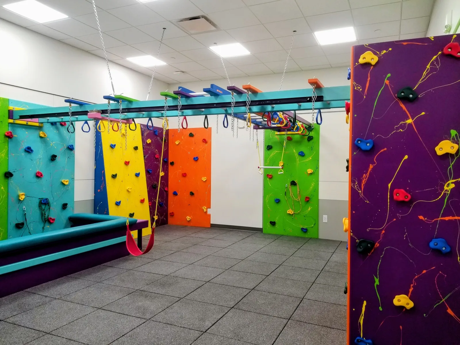 Colorful climbing walls, overhead rings, and a foam pit in a tiled hospital sensory gym