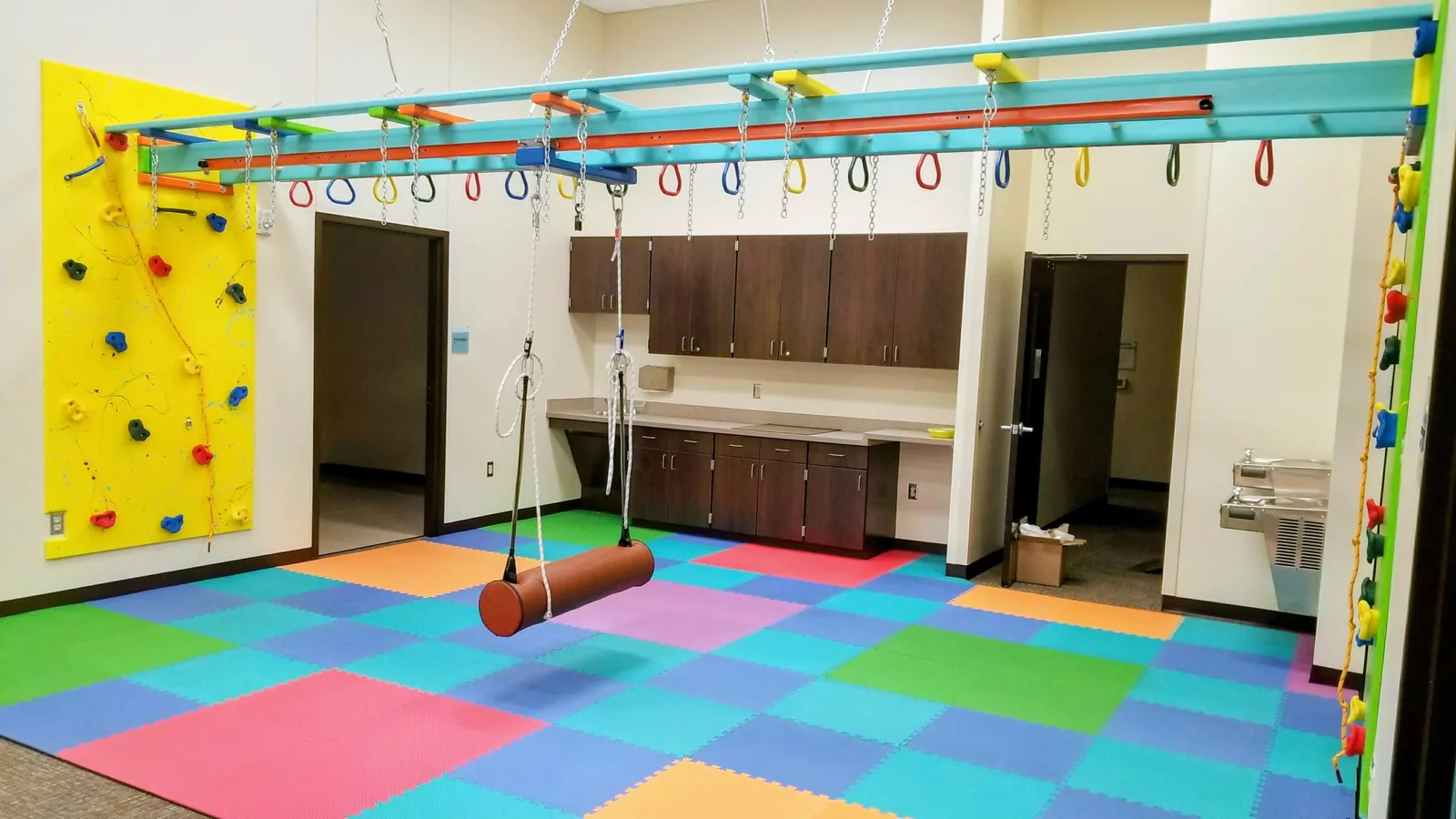Long sensory gym with colorful floor mats, a suspended bolster swing, and an overhead ring track