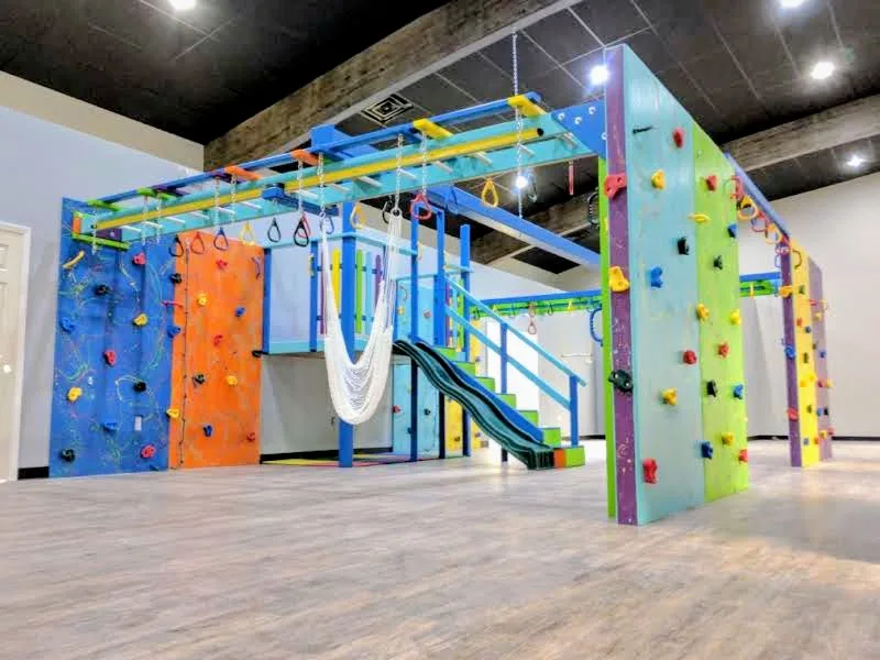 Large sensory gym with colorful climbing panels, overhead monkey bars, a white hammock, and a green slide
