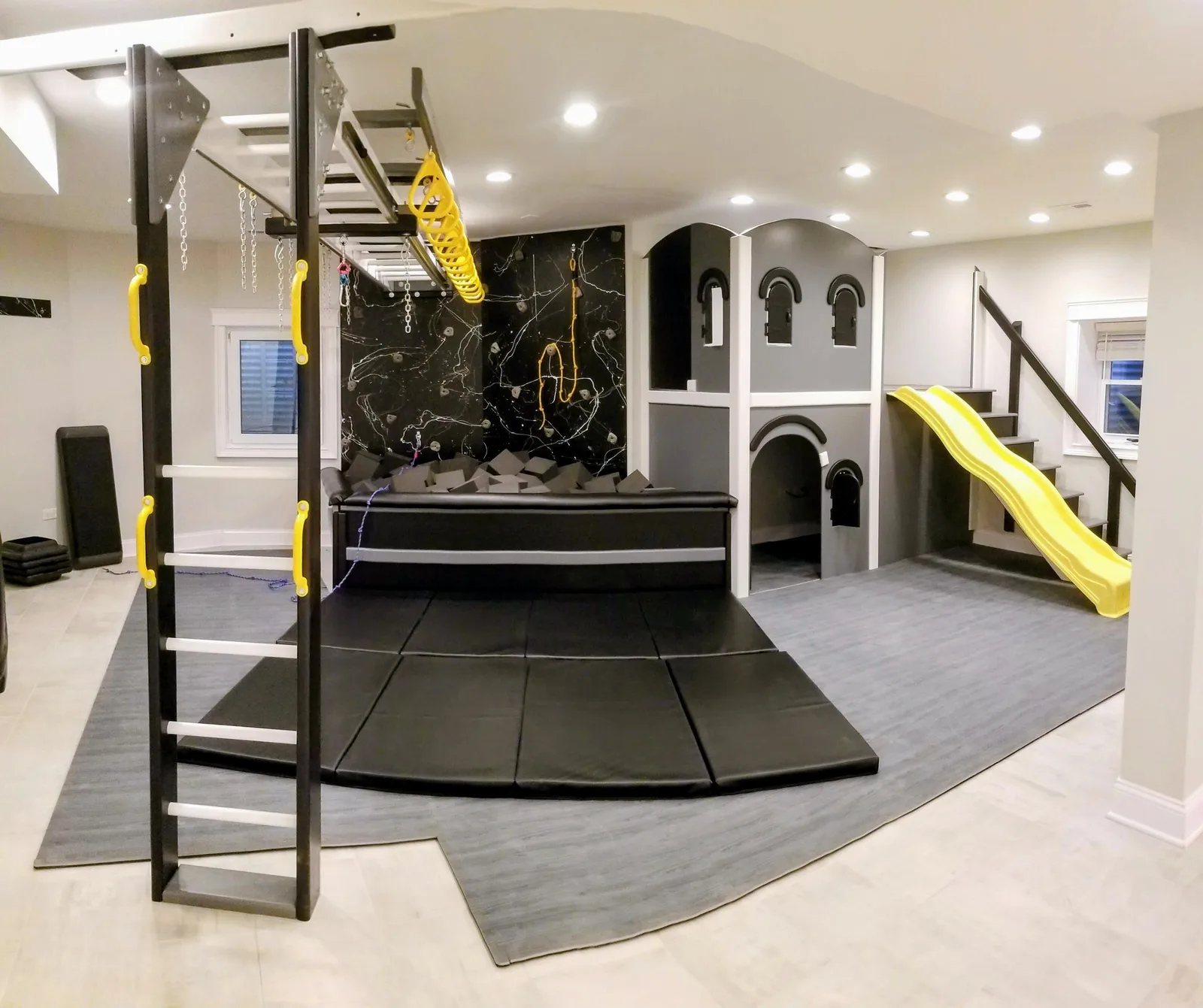 Dark residential sensory gym with yellow overhead bars, a yellow slide, padded flooring, and a castle structure